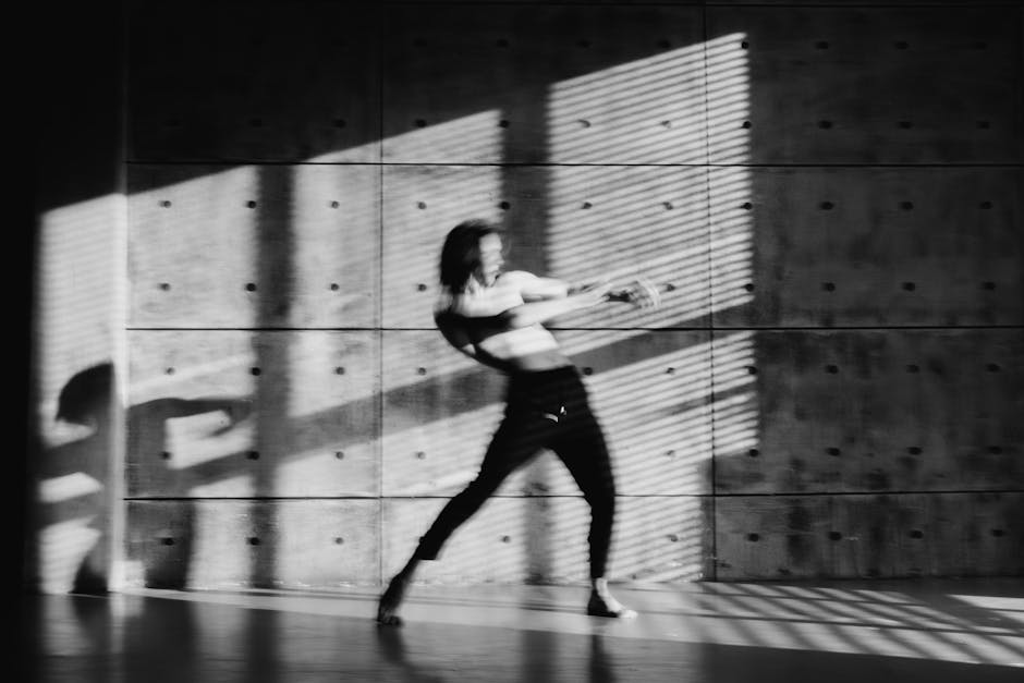 Grayscale image of a dancer in motion, casting a dramatic shadow in an indoor setting.